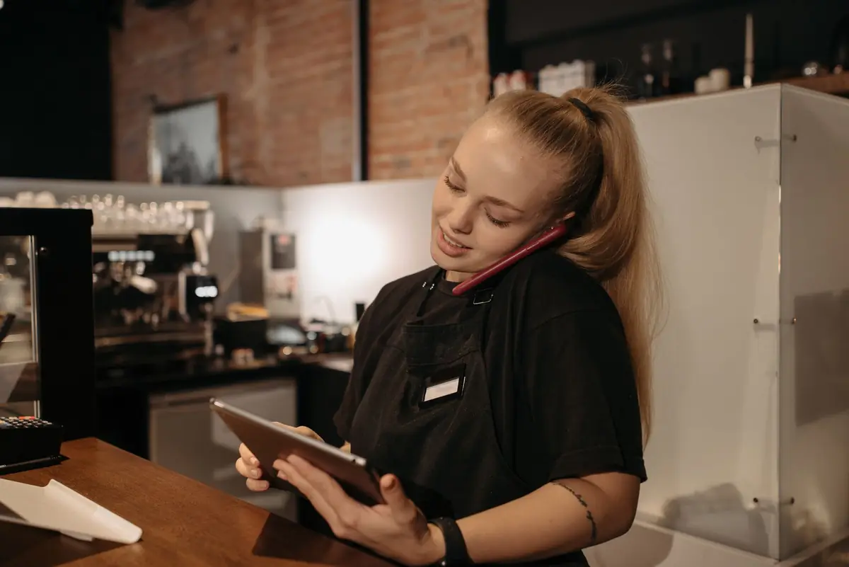 Een vrouw achter de toonbank van een café, aan de telefoon met een tablet in haar hand