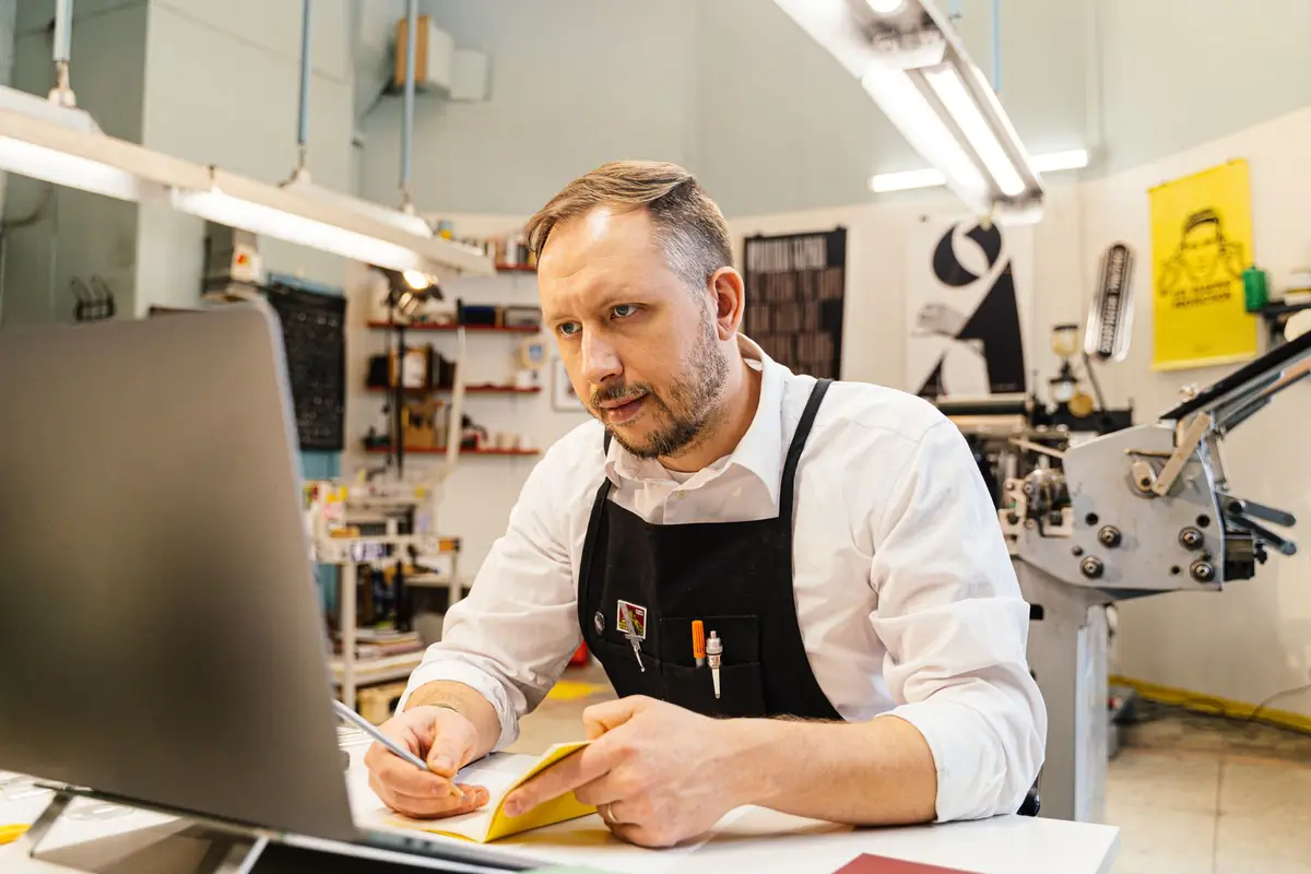 Een man met een schort aan zijn laptop in een drukkerij